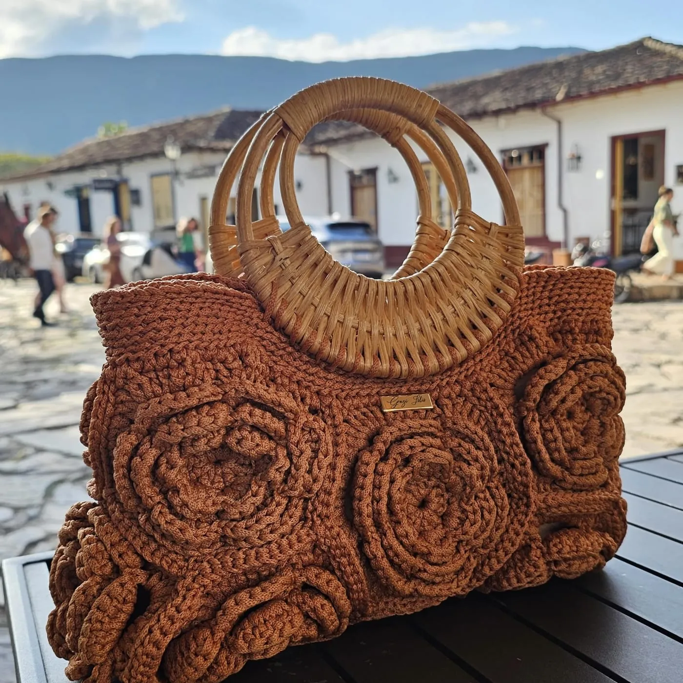 Bolsa Floral Elegância caramelo, com alças de rattan, numa rua de cidade histórica